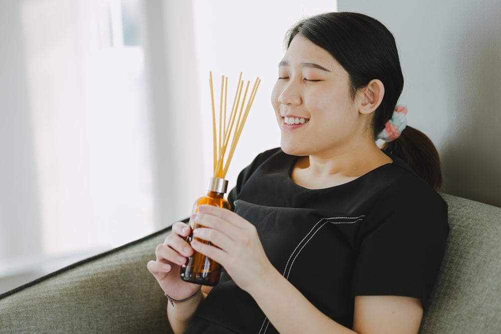 一位年輕女士正舒適地坐在沙發上，手持空氣清新劑擴香瓶，沉醉於香氣之中。這類產品雖營造愉悅感，但其化學成分亦可能對敏感體質構成風險。