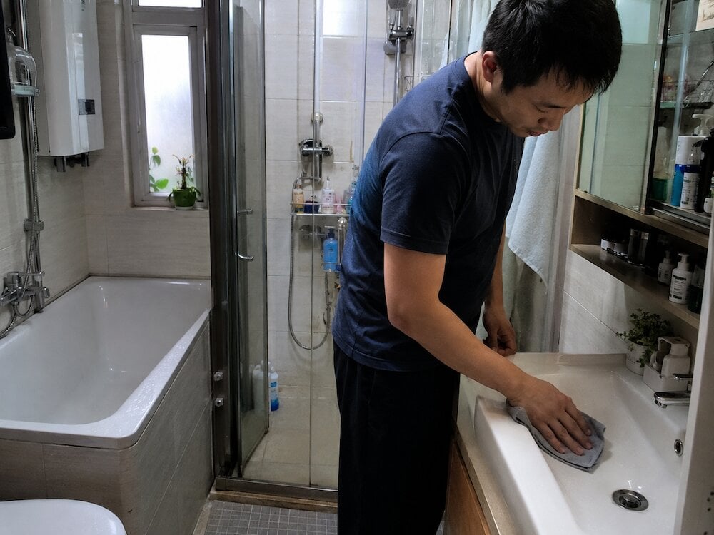 防水膠 一名穿海軍藍襯衫男子用抹布清潔浴室洗手盤,浴缸和淋浴間邊緣均勻塗上防水膠,旁有盥洗用品架.